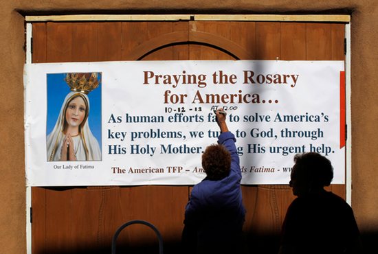 Two women hang a sign advertising "Praying the Rosary for America ..." outside the historic St. Francis Church in Taos, N.M., Oct. 5. CNS photo/Brian Snyder, Reuters